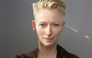 Actress Tilda Swinton is pictured in headshot, barely smiling, with short, backswept, blond hair and intense, blue-gray eyes.